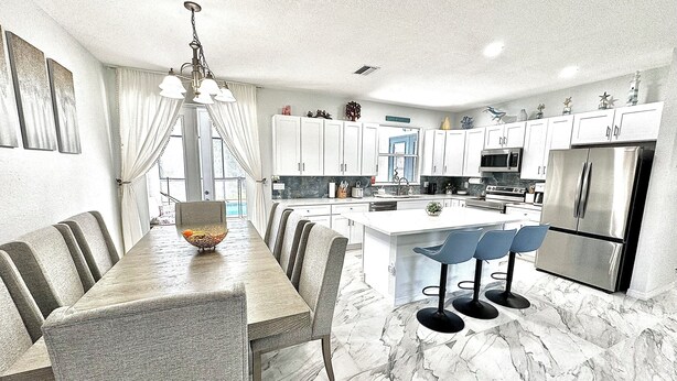 A modern kitchen with a marble floor, white cabinets, a central island, and a dining area with grey upholstered chairs. - New-North Port Florida -Heated pool - 5 BED (North Port)