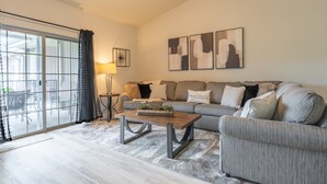 Ein Wohnzimmer mit grauem Sofa, einem Holztisch und einem großen Fenster mit Vorhängen.