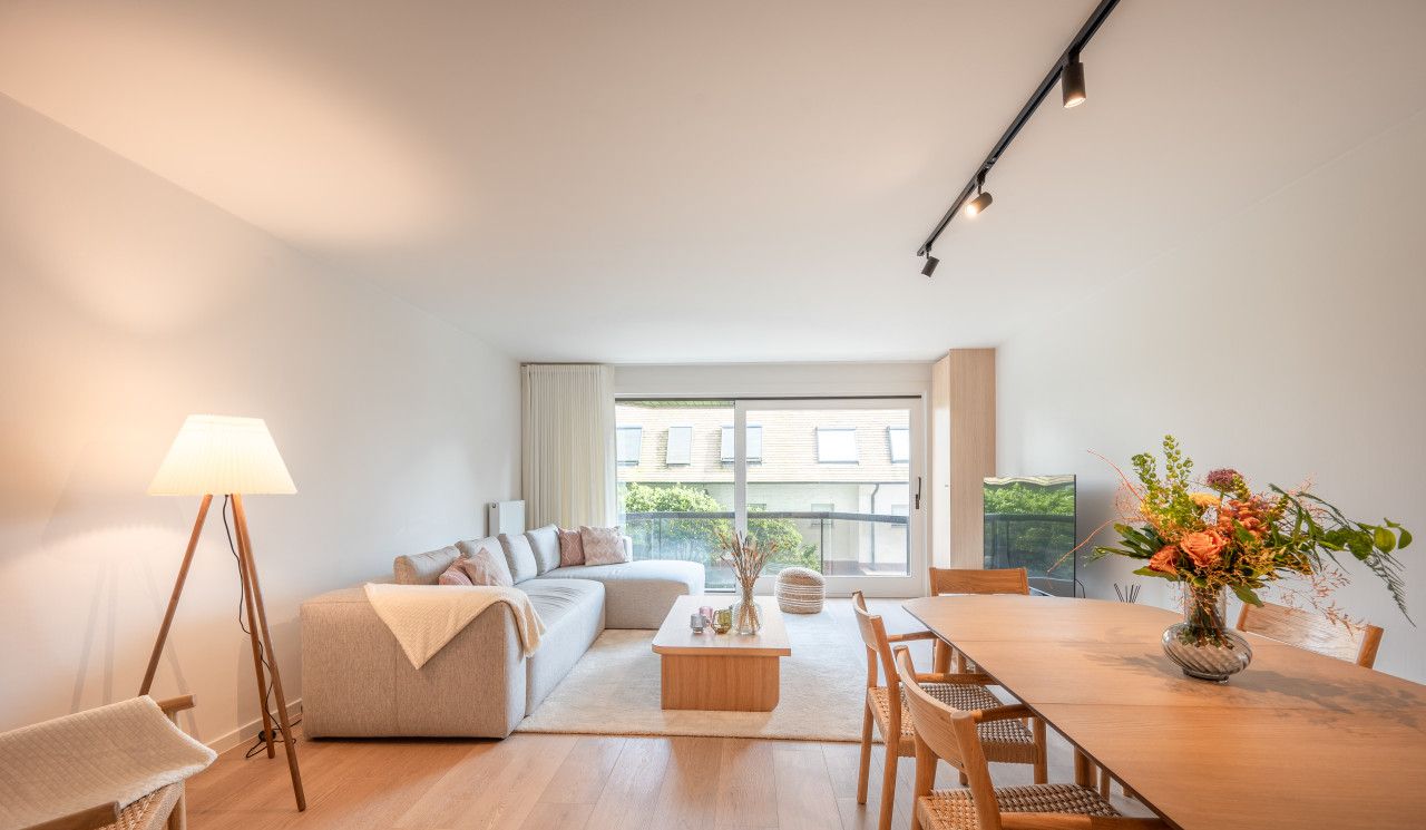 A modern living room with a sofa, coffee table, and dining table. There is a lamp, a vase with flowers, and a view of the outside through large windows.