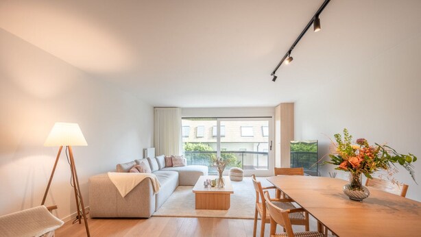 A modern living room with a sofa, coffee table, and dining table. There is a lamp, a vase with flowers, and a view of the outside through large windows.