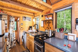 A compact kitchen with a stove, sink, and a window overlooking a forest. - Tiny bungalow nestled in the towering pines (Nevis)