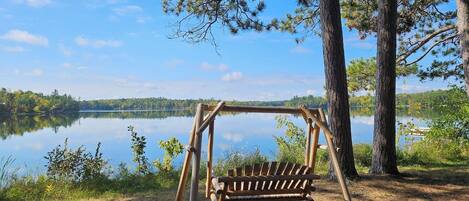 A wooden swing set with a bench, surrounded by trees and a lake.