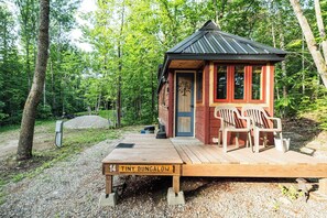 A tiny wooden bungalow with a porch, two chairs, and a sign indicating its number. - Tiny bungalow nestled in the towering pines (Nevis)