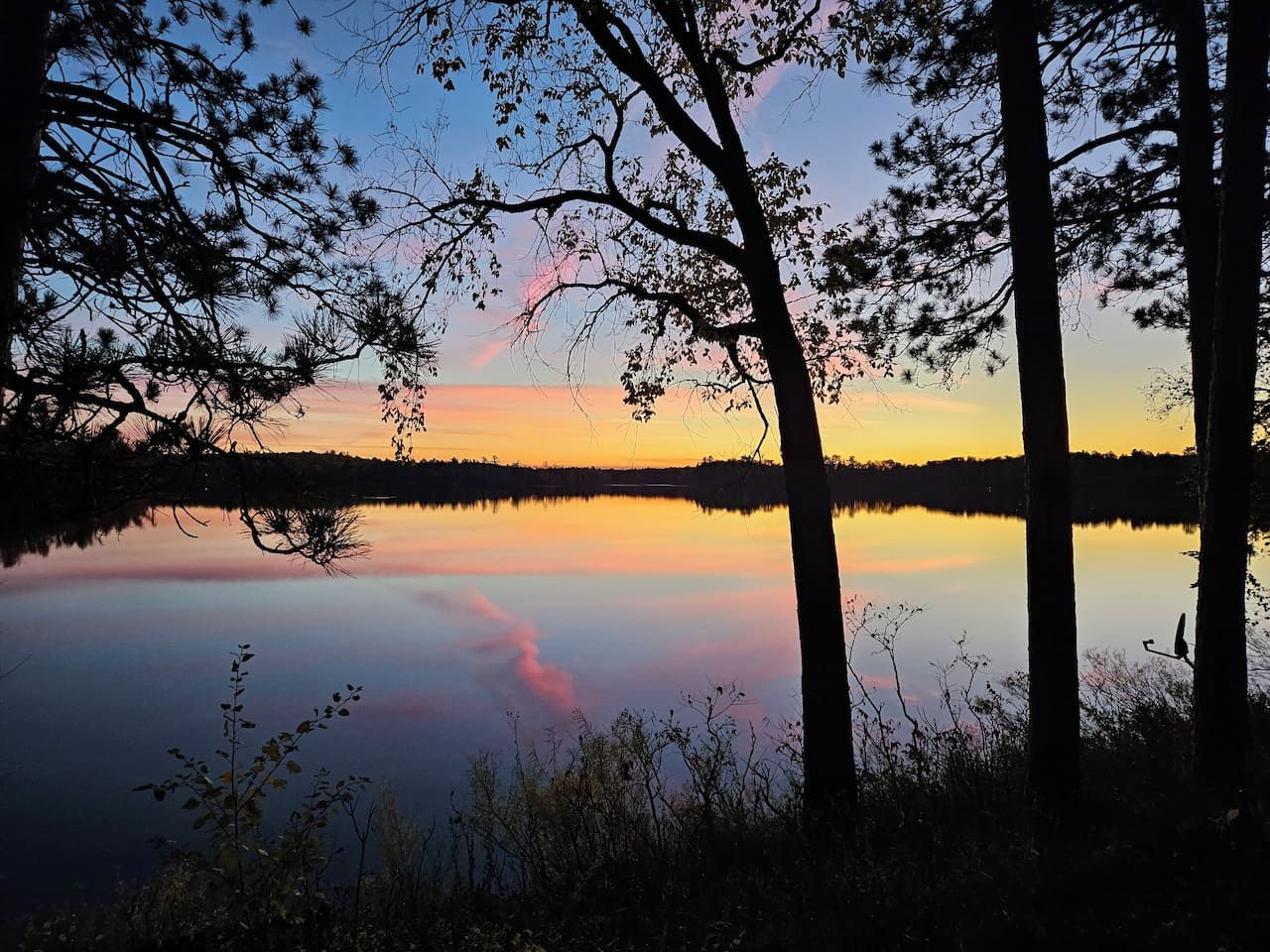 Un lac paisible, bordé d’arbres dont le reflet se mire dans le ciel au coucher du soleil.