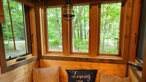 L'intérieur d'un chalet en bois, avec un banc, un coussin orné de l'inscription « Living The Dream » et une vue sur une forêt, visible à travers de grandes fenêtres.