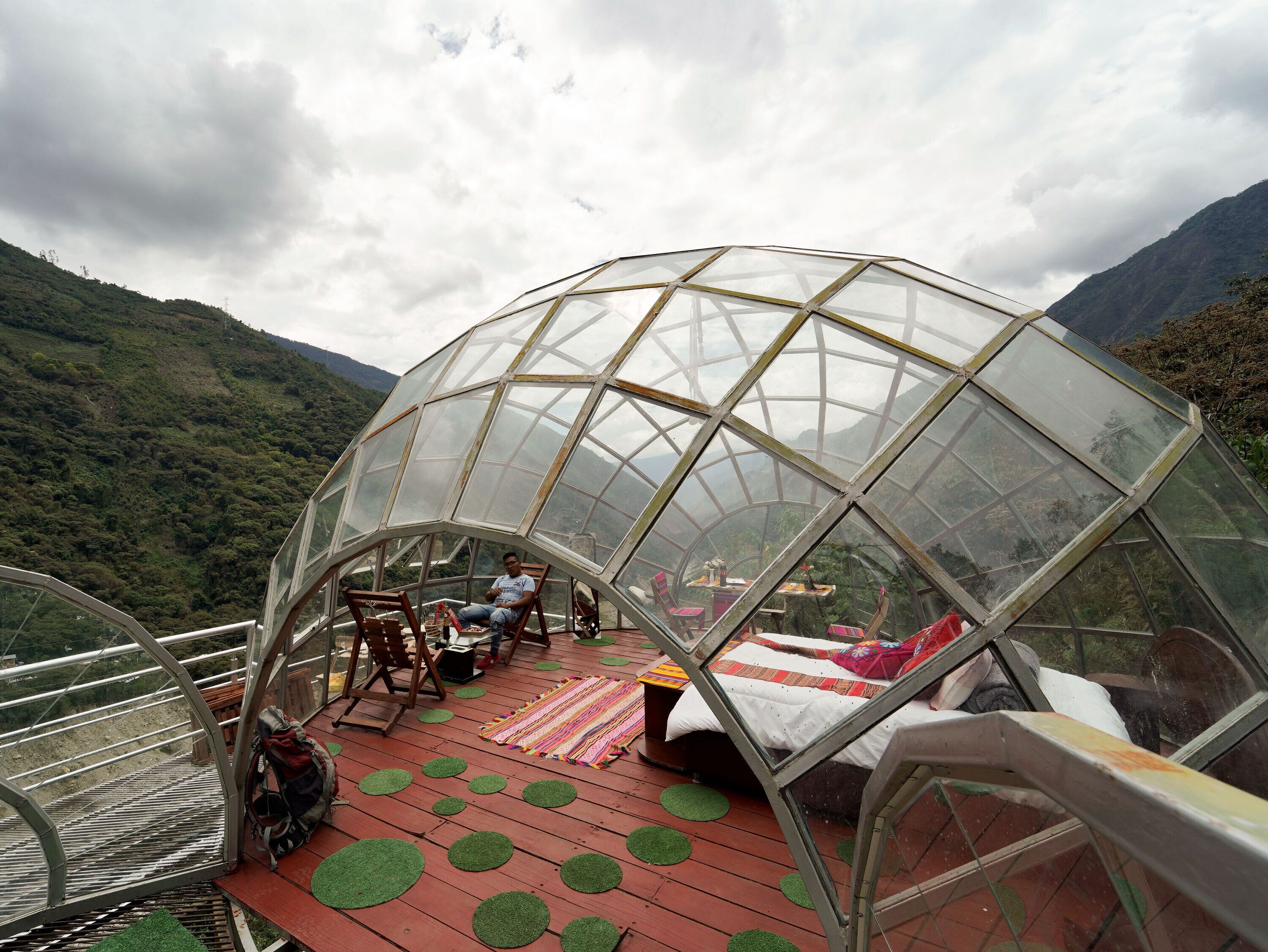 Estructura con cúpula de vidrio que contiene una cama y zona de estar, rodeada por una terraza roja con esteras verdes, con montañas al fondo.