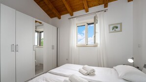 Un dormitorio con cama, cortinas blancas, ventana, lámpara de noche y armario.