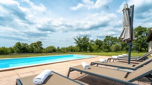Zona junto a la piscina con sillones de descanso y una sombrilla, rodeada de vegetación y un cielo despejado.