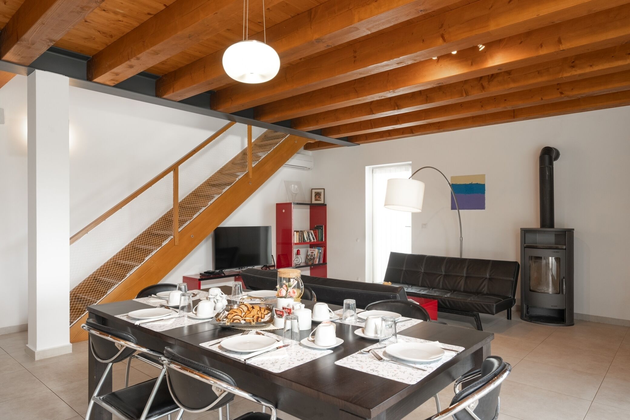Zona pranzo con tavolo di legno apparecchiato per quattro persone, una scala, un divano nero, un televisore e un camino a legna.