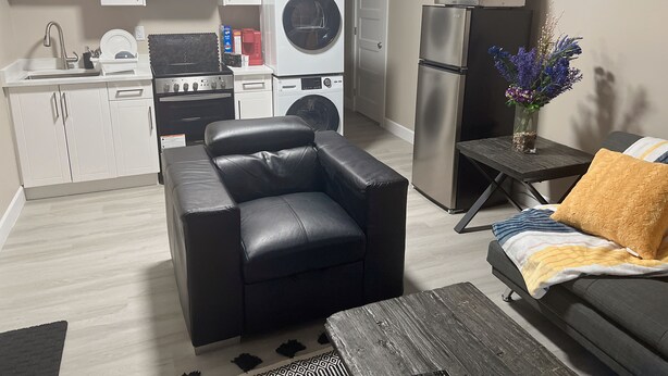 Un espace de vie compact comprenant un coin cuisine, un canapé en cuir noir, une table basse en bois et une petite table d’appoint agrémentée d’un vase de fleurs.