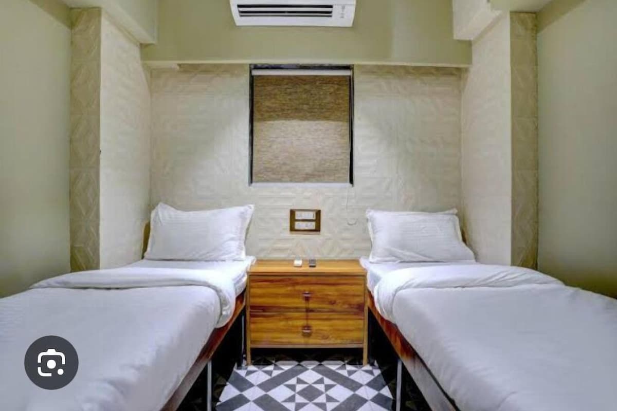 A small, clean room with two single beds, a wooden nightstand, and a wall-mounted air conditioning unit.