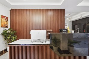 A reception desk with a computer, a brown wooden panel wall, a potted plant, and a painting on the wall. - Hotel Amahi Inn Sector 30 (Gurugram)