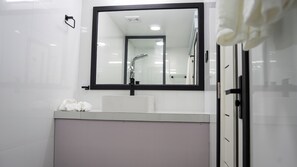Un baño moderno con un lavamanos blanco, un espejo grande y una ducha visible en el reflejo.