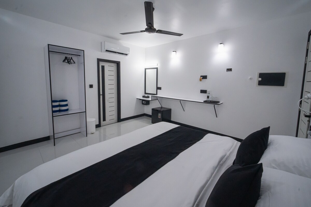 Una habitación de hotel moderna con una cama grande, un ventilador de techo, un armario, una mesita de noche pequeña con una lámpara y un espejo en la pared.