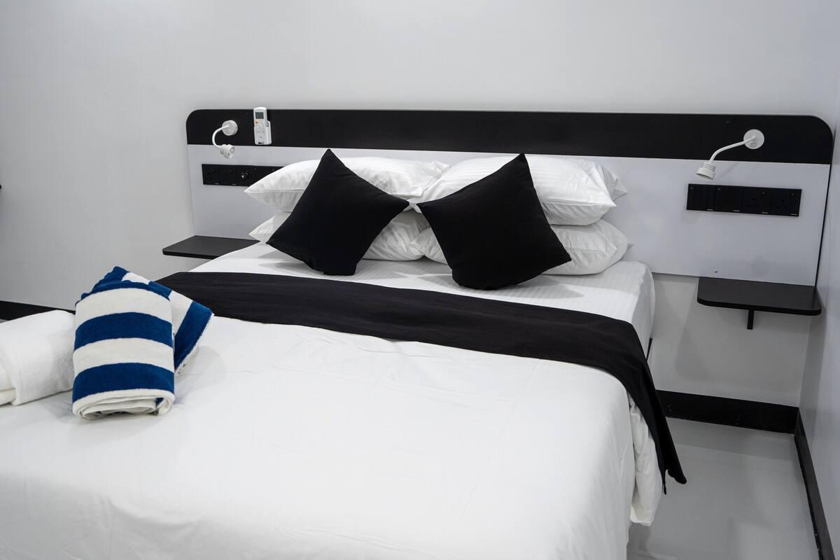 A neatly made bed with white and black bedding, a striped towel, and a black pillow. There are two bedside tables with a phone and a lamp on each.