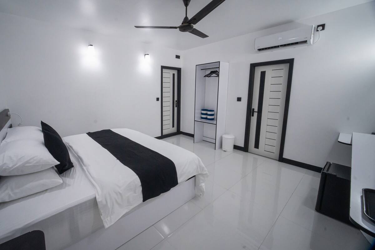 A modern hotel room with a bed, bedside table, ceiling fan, air conditioning unit, and a wardrobe.