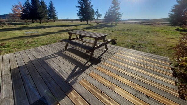 A wooden deck with a picnic table in a grassy field with trees in the background.
