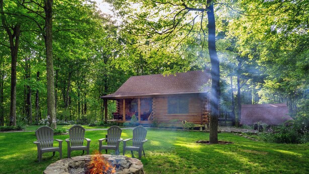 Una cabaña con un pozo de fuego de piedra rodeado de sillas Adirondack, ubicada en un bosque frondoso.