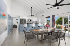 A modern kitchen with white cabinets, a central island, and a dining area with wicker chairs and a wooden table. - The Corktree Villa: 3 Bedroom, by Olympic Residences (Turks Islands)
