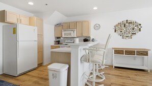 A compact kitchen with a refrigerator, microwave, and a sink. There is a white trash bin and a bar stool with a white chair. A clock is on the wall. - Blue Oasis - Two Pools, Beach, & More! (Osage Beach)