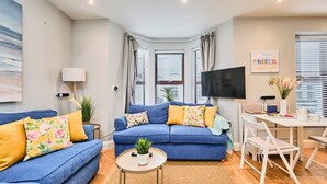 Un salon moderne avec un canapé bleu, une petite table basse ronde, un téléviseur et un coin repas comprenant une table et des chaises blanches.