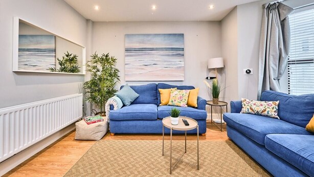 Un salon moderne avec un canapé bleu, une table basse et deux petites tables d’appoint. Deux grandes peintures ornent le mur, ainsi qu’une lampe sur pied et une plante en pot.