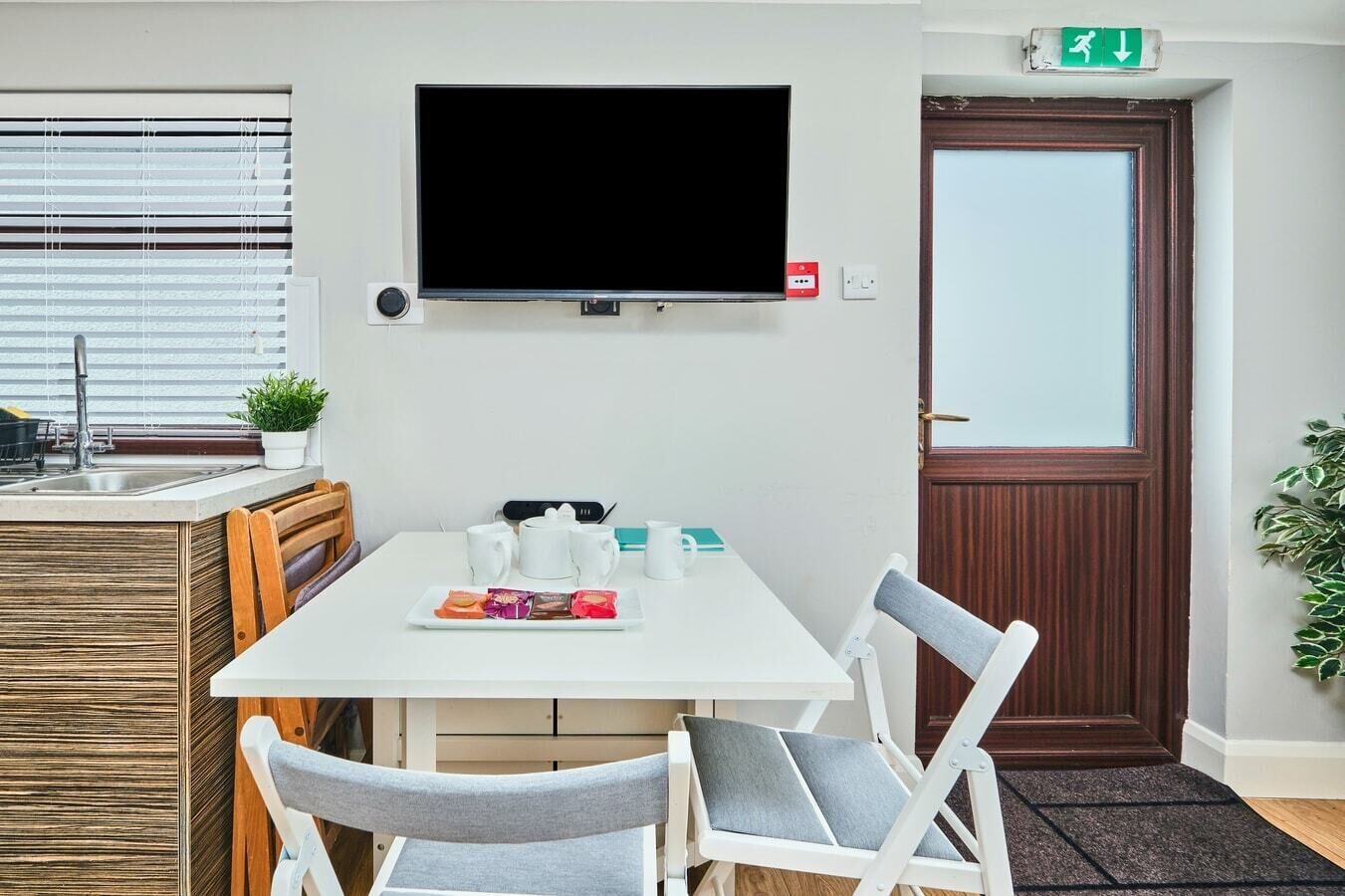 A hotel room with a white dining table, two chairs, a flat-screen TV mounted on the wall, a small kitchen area with a sink and a potted plant, and a door with an emergency exit sign.