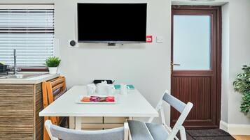 A hotel room with a white dining table, two chairs, a flat-screen TV mounted on the wall, a small kitchen area with a sink and a potted plant, and a door with an emergency exit sign.