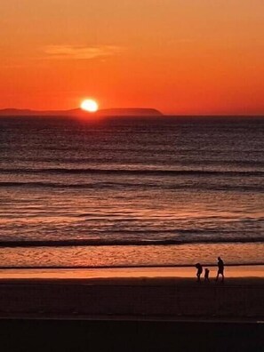 Solnedgang over havet med en strand og folk, der går langs kysten.