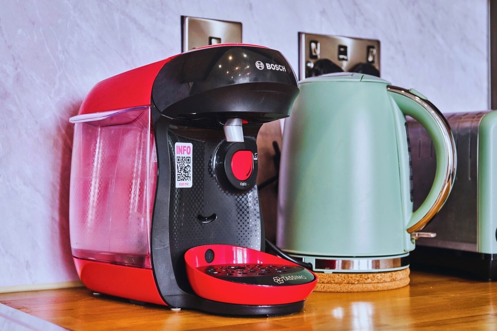 Una cafetera roja y negra con un código QR y un hervidor verde sobre una superficie de madera.