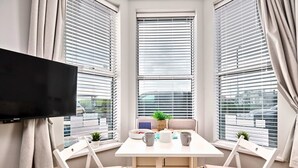 Un comedor con una mesa y sillas, un televisor de pantalla plana en la pared y ventanas grandes con persianas.