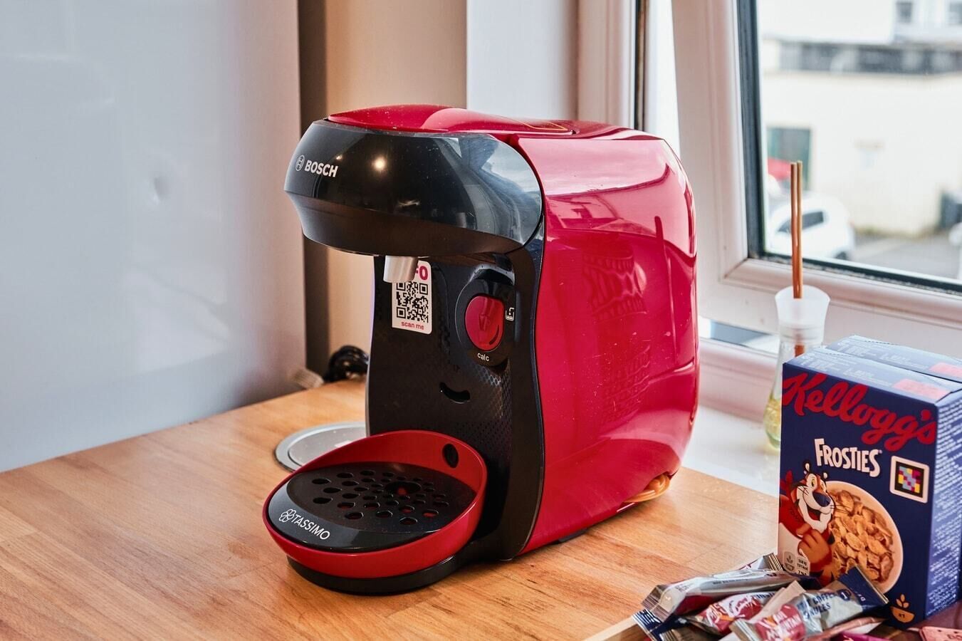 A red and black coffee machine with a QR code, a bowl, and a box of Kellogg's Frosties cereal on a wooden table near a window.