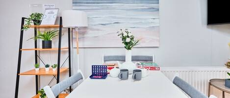 A modern dining area with a white table, grey upholstered chairs, a bookshelf with plants, a painting of the ocean, and a white radiator.