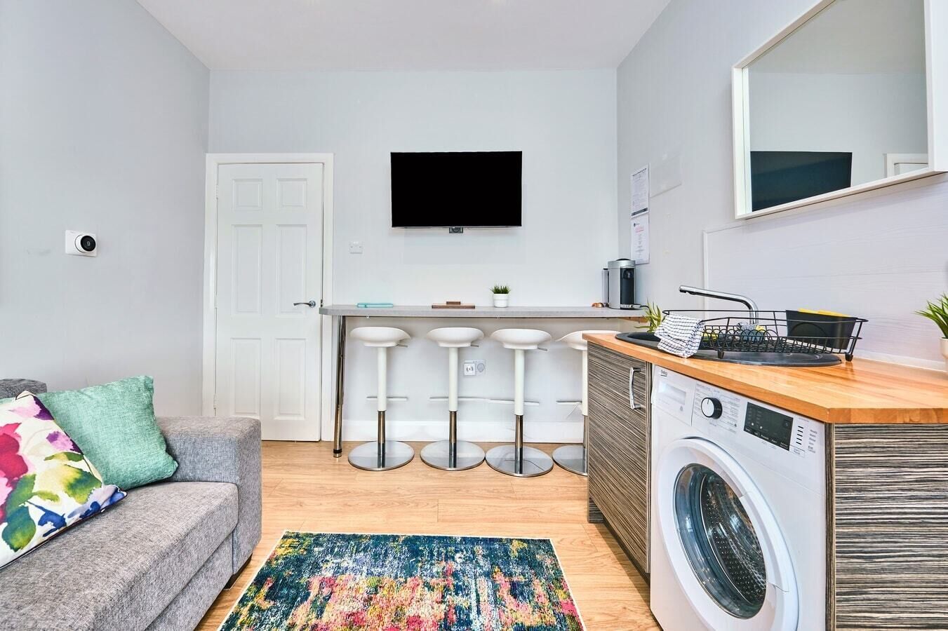 A compact living space with a kitchenette, a washing machine, a sofa with colorful cushions, and a bar stool area.