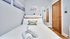 Una cama bien tendida con una almohada que dice 'nuestro lugar feliz', una mesita de noche con una lámpara y una puerta de madera.