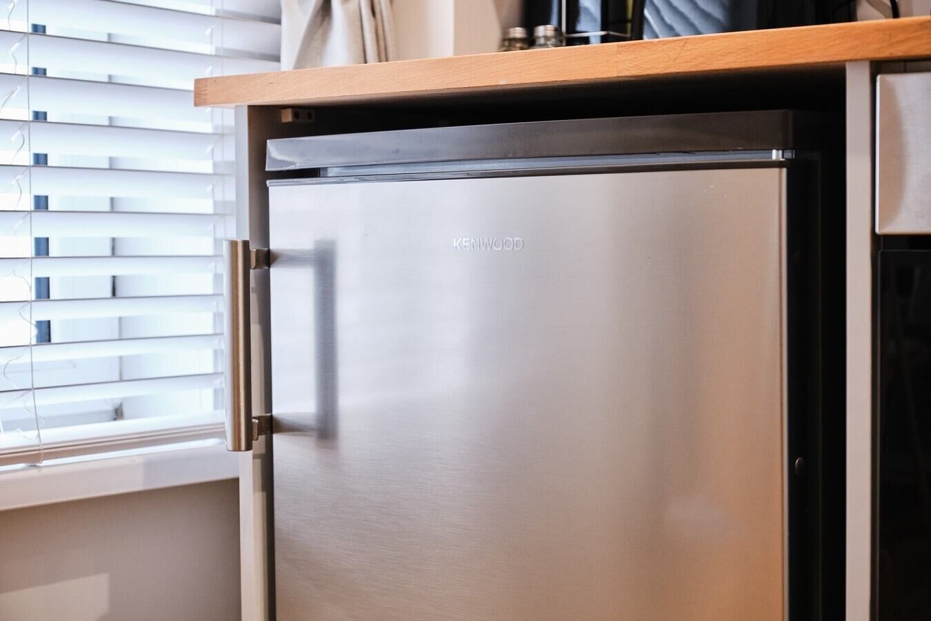 Un refrigerador Kenwood en una cocina con encimera de madera y una ventana con persianas.