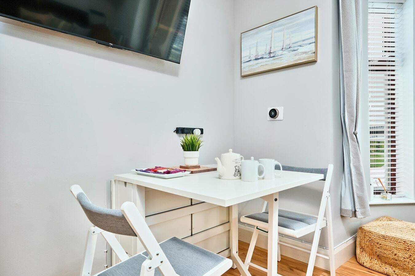 A small, modern room with a white table, two chairs, a TV mounted on the wall, a painting above the table, a potted plant, and a window with blinds.