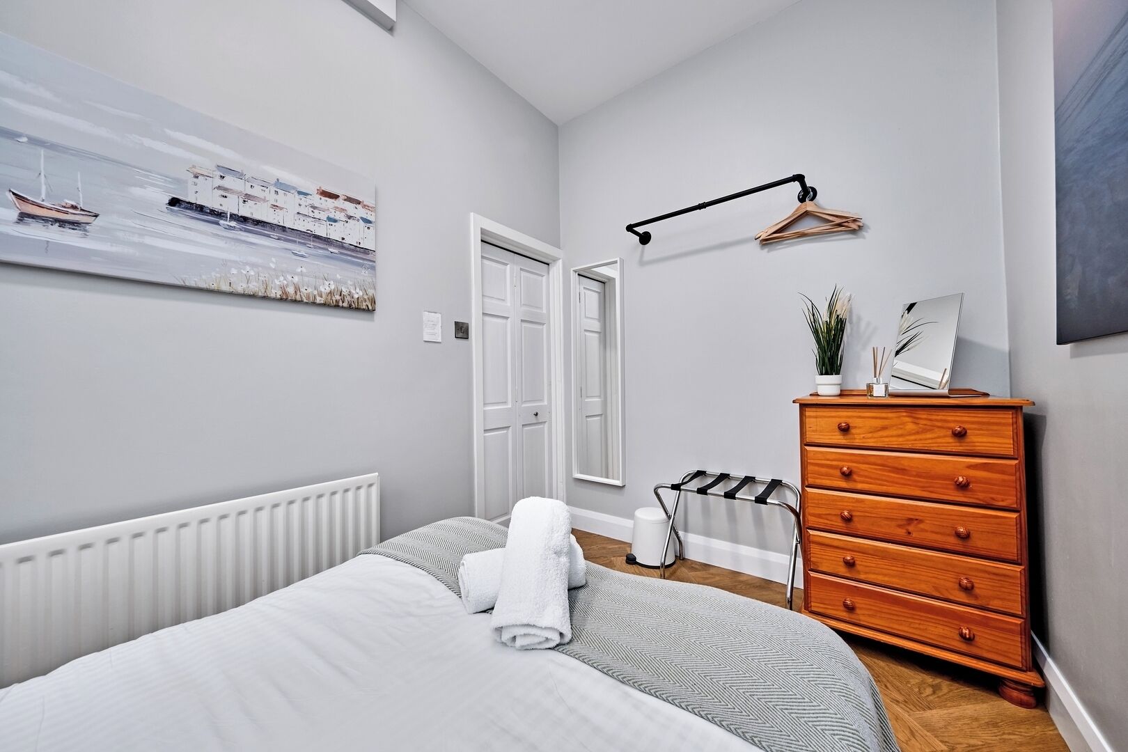 Ein Schlafzimmer mit Bett, Kommode, Heizkörper, einer Tür und einem an der Wand befestigten Handtuchhalter.