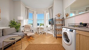 A modern living room with a sofa, armchair, coffee table, and a TV. There's a dining area with a table and chairs, and a kitchen with a washing machine.