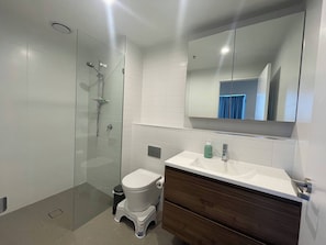 A modern bathroom with a glass-enclosed shower, a white toilet, a wooden vanity with a white sink, and a large mirror above the vanity. - 2BR Apartment with Pool and Gym (Phillip)