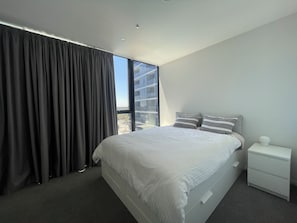 A modern bedroom with a large bed, a nightstand, and a view of a building through a window. - 2BR Apartment with Pool and Gym (Phillip)