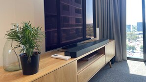Uma sala de estar moderna com uma grande televisão de tela plana na parede, um móvel de entretenimento de madeira abaixo, uma planta em vaso e vista para a cidade através de uma grande janela.