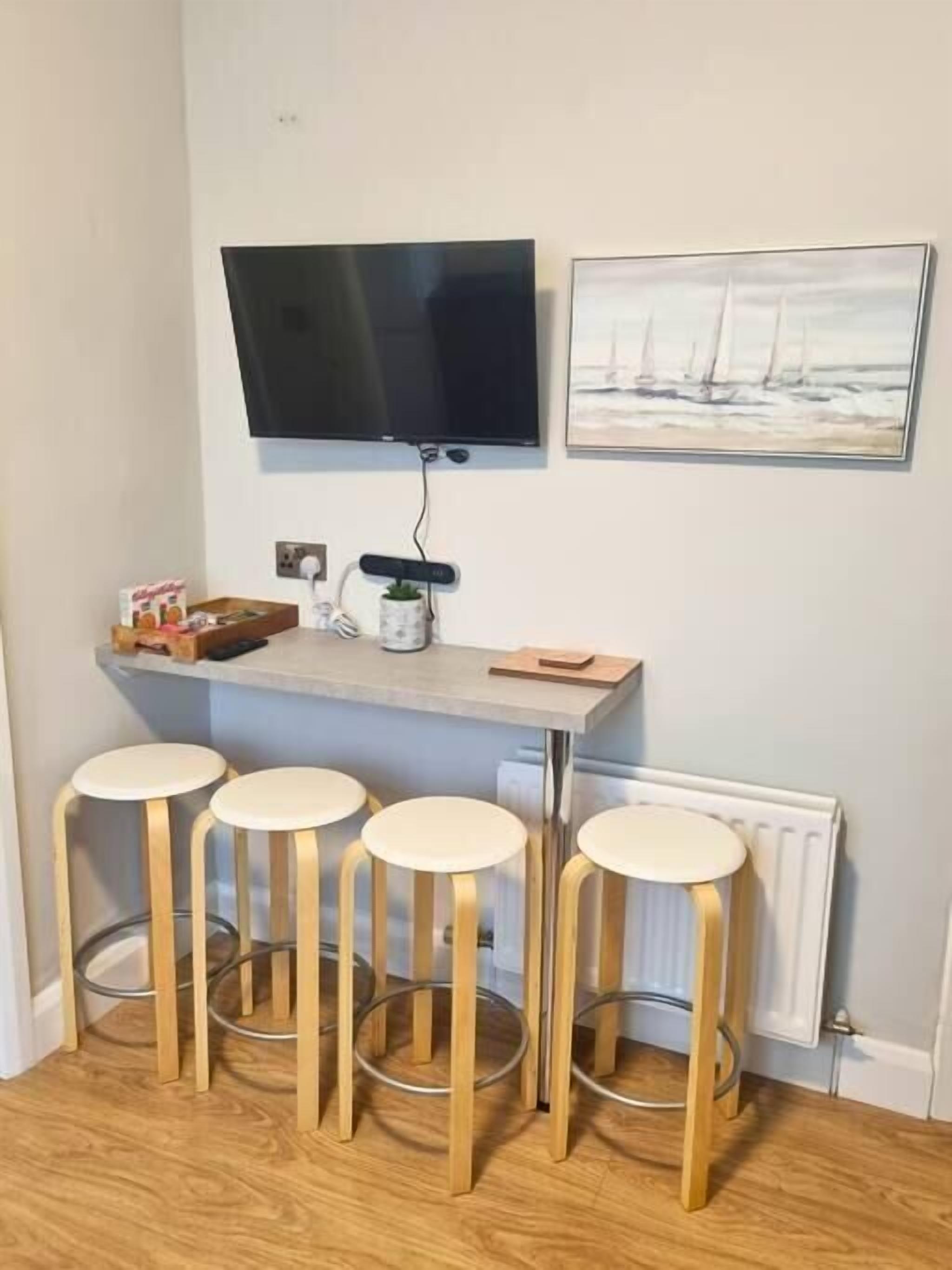 A small kitchen area with a TV mounted above a counter, a painting on the wall, and three bar stools.