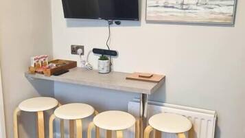 A small kitchen area with a TV mounted above a counter, a painting on the wall, and three bar stools.