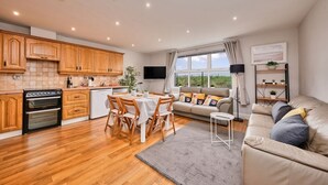 A modern kitchen with wooden cabinets, a dining table with chairs, a sofa, a TV, and a window with a view. - Spacious Bright Apt Walk to the Beach (Portrush)