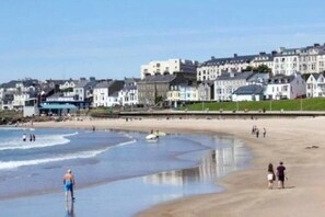 A beach with people walking and standing, a clear sky, and buildings in the background. - Spacious Bright Apt Walk to the Beach (Portrush)