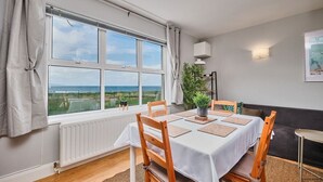 A dining area with a table set for four, large windows with a view of the sea, a radiator, and a plant on the windowsill. - Panoramic Sea View Beach Apartment (Portrush)