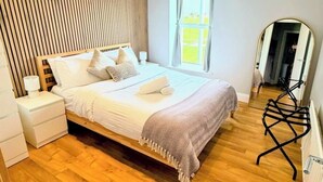 Une chambre avec un mur recouvert de lambris en bois, un lit avec du linge de lit blanc, une table de chevet, une chaise et une fenêtre avec des rideaux.