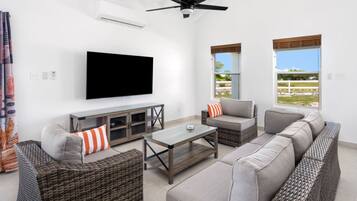 A modern living room with a flat-screen TV, wicker furniture, and large windows offering a view of the outdoors.