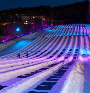 Een met sneeuw bedekte helling met een reeks parallelle lijnen, mogelijk bedoeld voor skiën of snowboarden, en een gebouw op de achtergrond.
